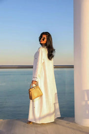 Woman wearing elegant white linen abaya with beige leaf embroidery standing by the sea holding a tan handbag.