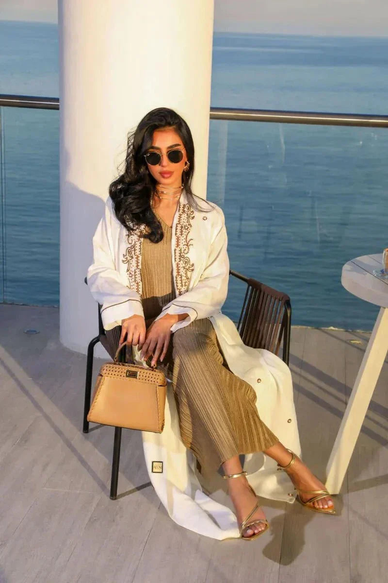 Woman wearing elegant white linen abaya with beige leaf embroidery standing by the sea holding a tan handbag.