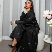 Woman wearing black satin abaya with beige embroidery and pleated chiffon cuffs sitting indoors