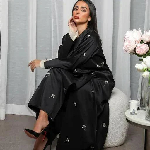 Woman wearing black satin abaya with beige embroidery and pleated chiffon cuffs sitting indoors