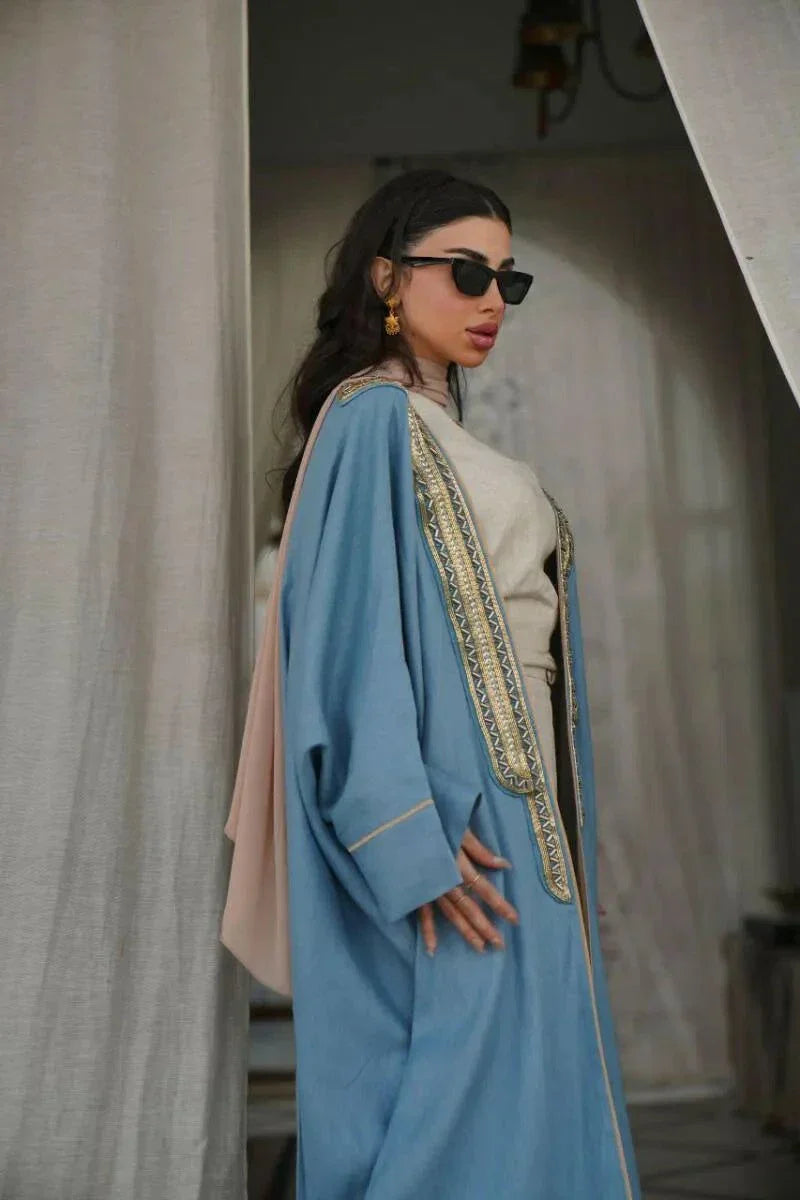 woman wearing soft blue linen bisht with golden embroidery and wide sleeves standing indoors