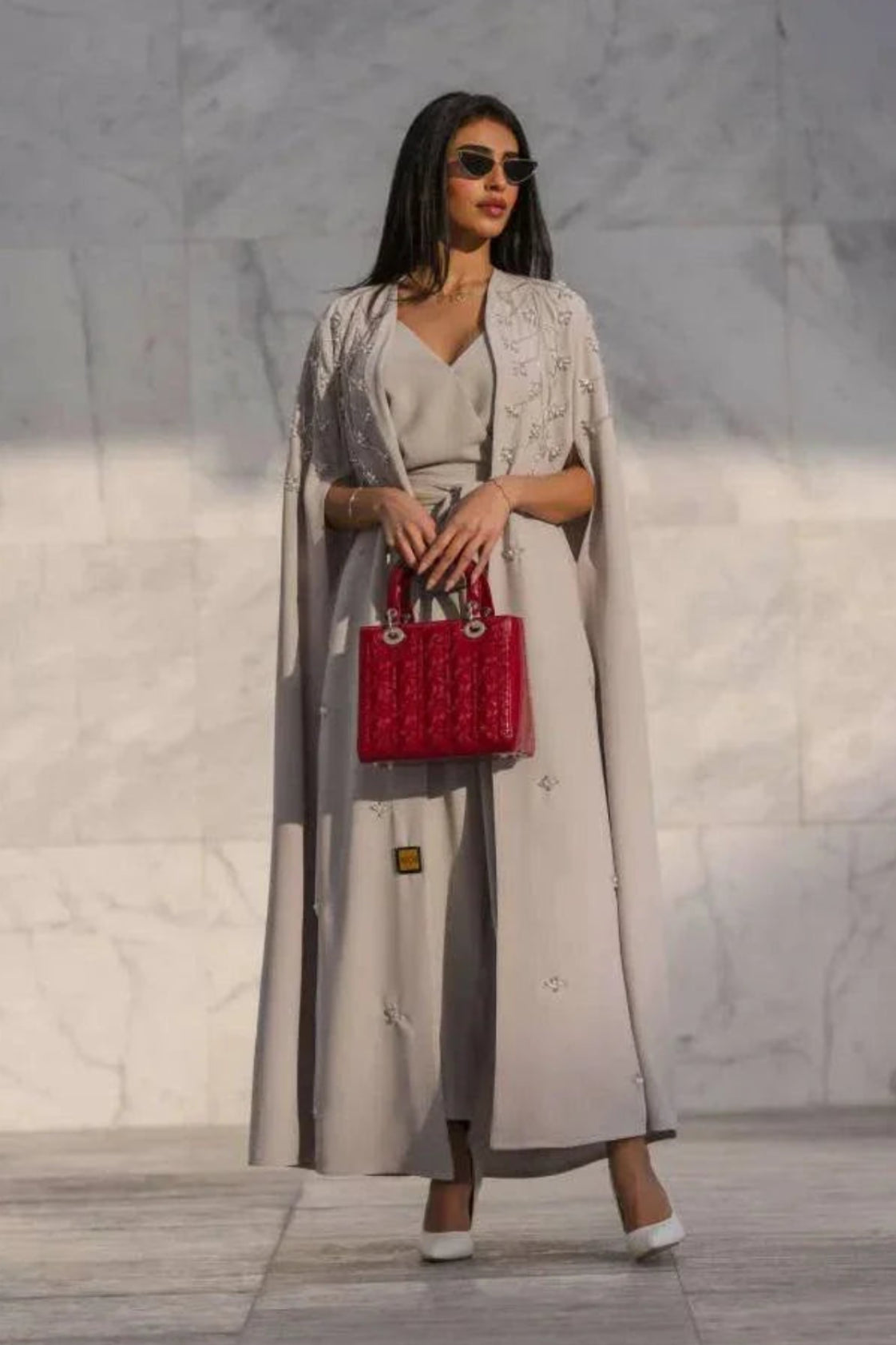 woman wearing light gray cape-style abaya with tonal embroidery holding red handbag against marble wall