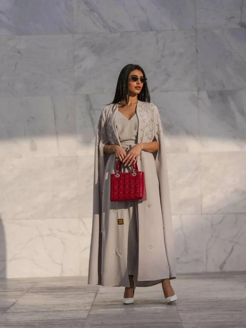 woman wearing light gray cape-style abaya with tonal embroidery holding red handbag against marble wall