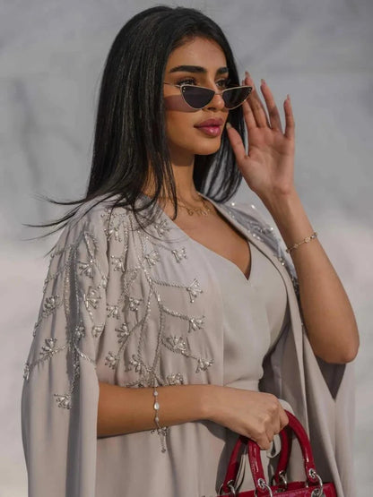 Light gray cape-style abaya with delicate tonal embroidery, made of fluid crepe fabric for a graceful silhouette, styled with sunglasses and a red handbag.