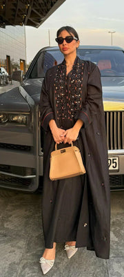 Woman wearing a black linen abaya with intricate orange embroidery, wide sleeves, and holding a beige handbag.