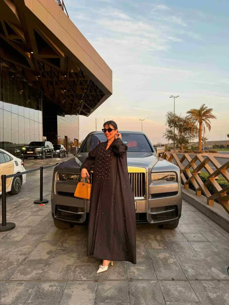 Woman wearing a black linen abaya with intricate orange embroidery, wide sleeves, and holding a beige handbag.