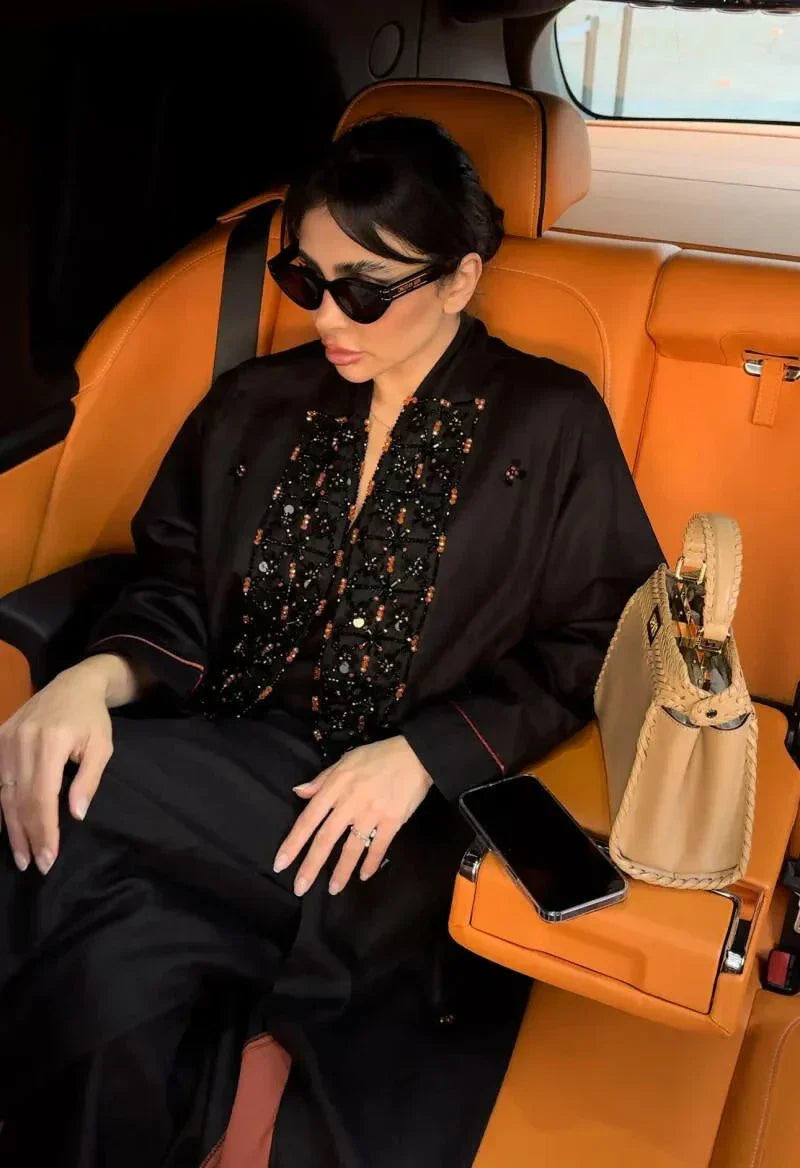 Woman wearing a black linen abaya with intricate orange embroidery, wide sleeves, and holding a beige handbag.