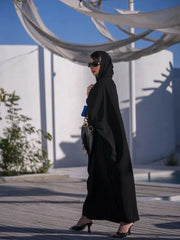 Woman wearing a flowy black Azraq abaya with blue embroidery and inner blue lining, paired with a matching sheila and black heels.