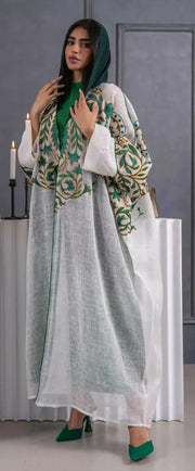 woman wearing lightweight white linen bisht with green and gold embroidery standing indoors holding a beige handbag