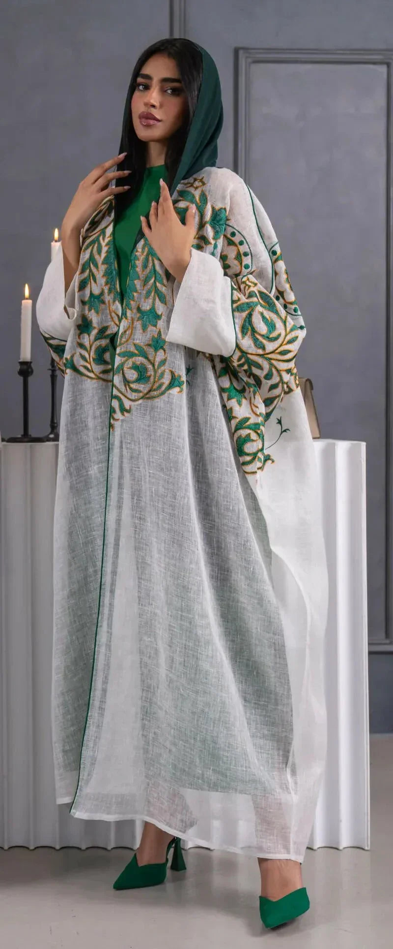 woman wearing lightweight white linen bisht with green and gold embroidery standing indoors holding a beige handbag
