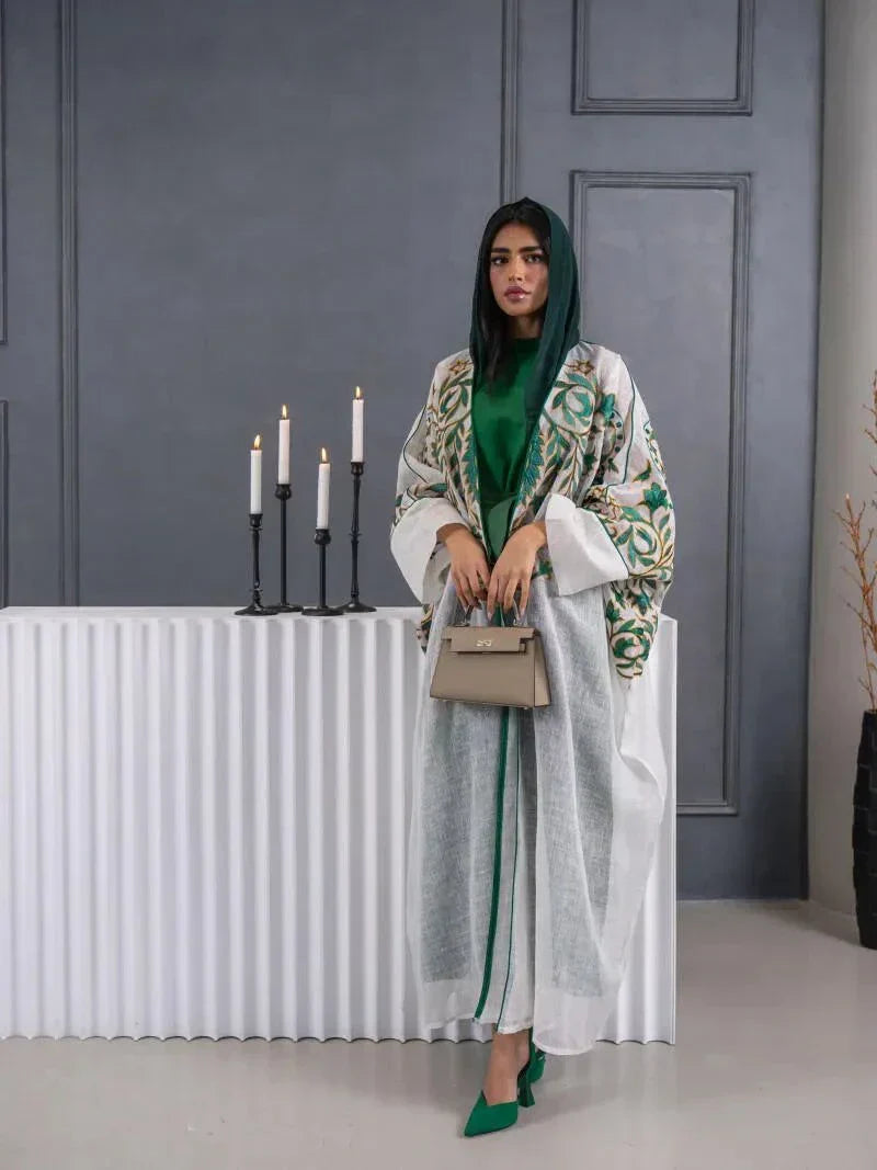 Woman wearing lightweight white linen bisht with green and gold embroidery over green dress holding beige handbag indoors.