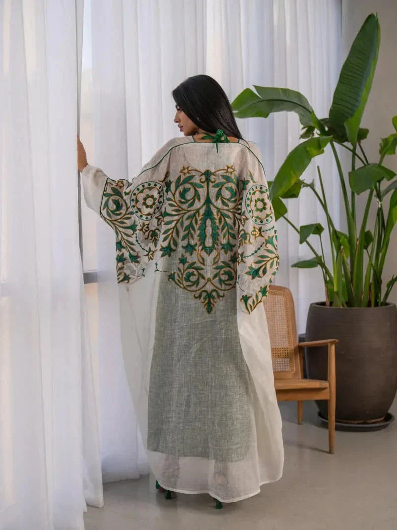 woman wearing lightweight white linen bisht with green and gold embroidery standing indoors holding a beige handbag