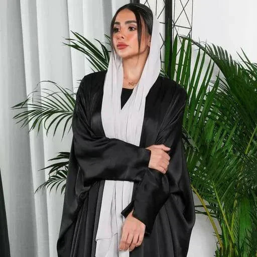 woman wearing flowy black bisht made of silky lightweight fabric standing indoors with green plants in background