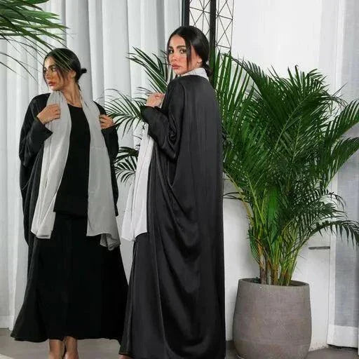 woman wearing flowy black bisht made of silky lightweight fabric standing indoors with green plants in background