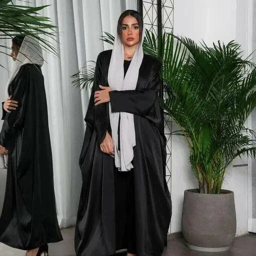 woman wearing flowy black bisht made of silky lightweight fabric standing indoors with green plants in background