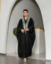 Woman wearing a luxurious black bisht with silver embroidery on the neckline and cuffs, standing in an archway.