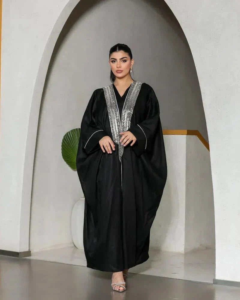 Woman wearing a luxurious black bisht with silver embroidery on the neckline and cuffs, standing in an archway.