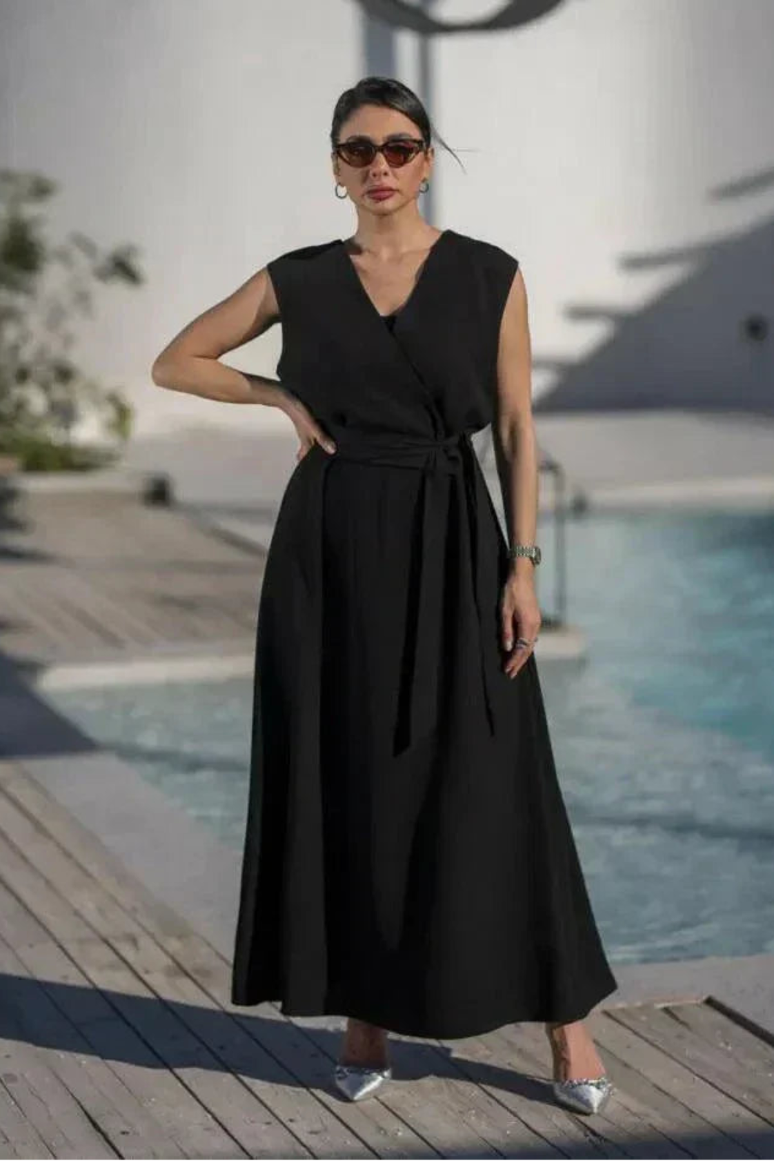 woman wearing sleeveless black wrap dress with waist tie standing by poolside