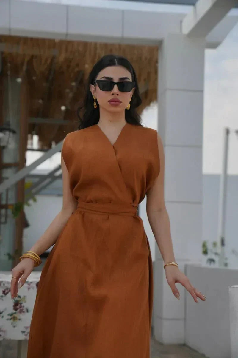 Woman wearing a sleeveless rust-toned linen dress with a V-neckline and waist belt standing outdoors under a pergola - Rustina