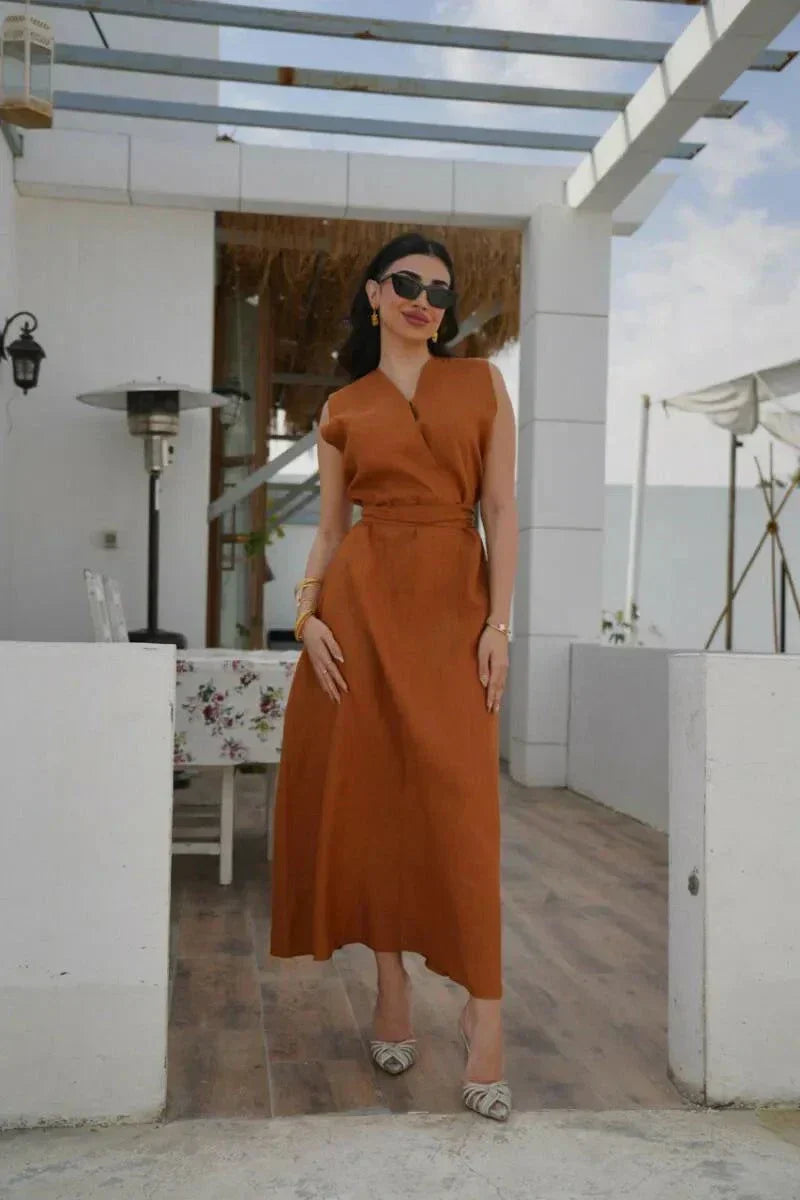 Woman wearing a sleeveless rust-toned linen dress with a V-neckline and waist belt standing outdoors under a pergola - Rustina