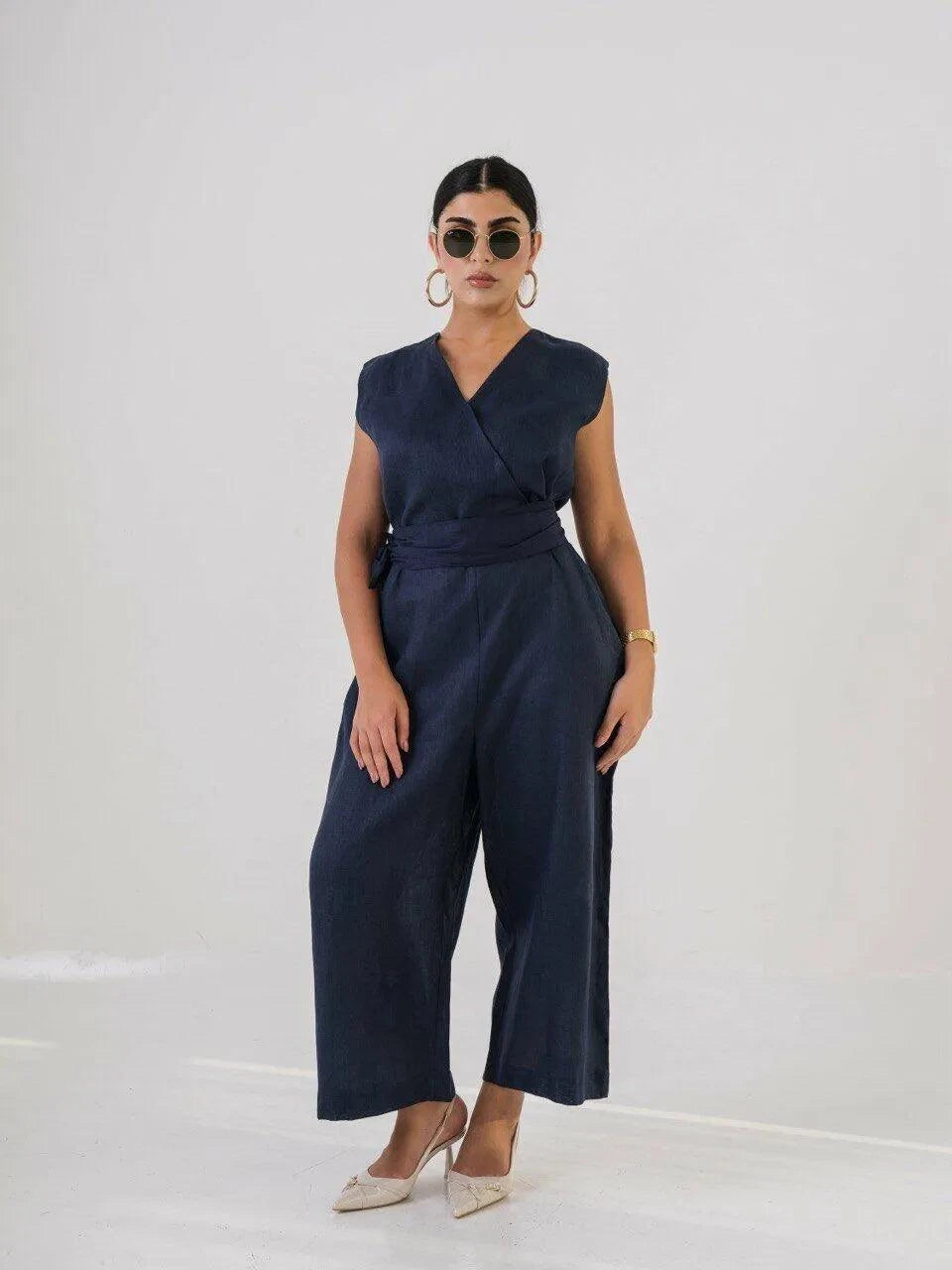 Woman wearing navy linen jumpsuit with V-neckline, fabric belt, and wide-leg pants, styled with sunglasses and heels