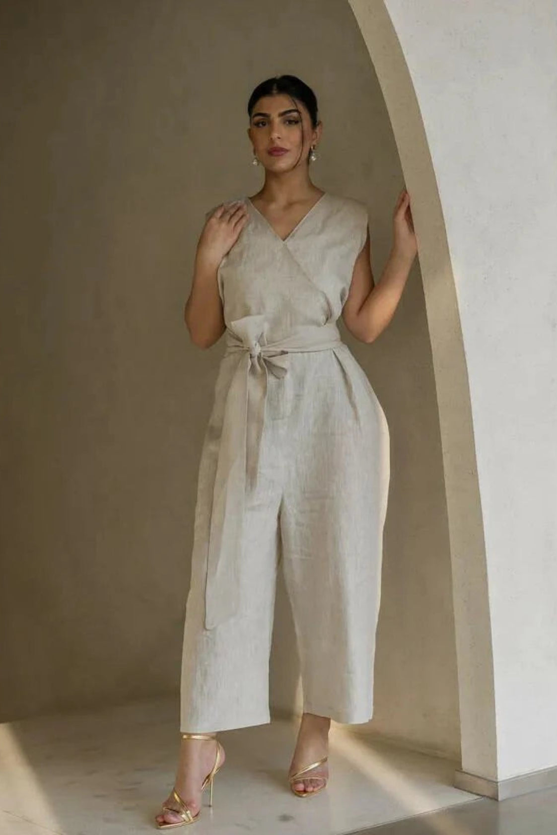 Woman wearing a beige linen jumpsuit with V-neckline, self-tie belt, and wide-leg silhouette standing by an archway
