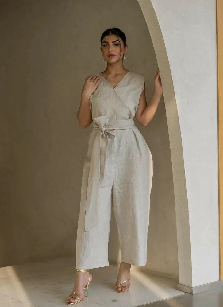 Woman wearing a beige linen jumpsuit with V-neckline, self-tie belt, and wide-leg silhouette standing by an archway