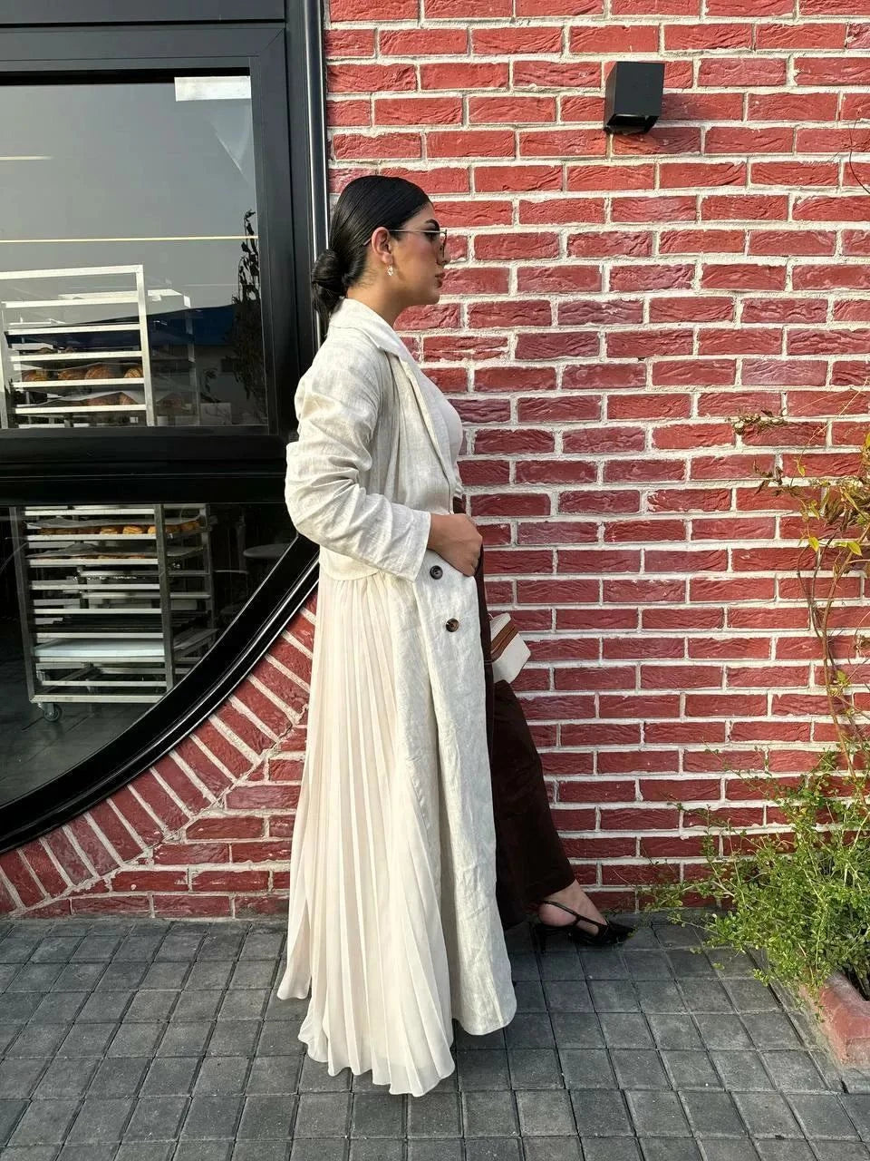 Woman wearing Whisper Beige linen abaya with pleated chiffon panels and double-breasted buttons, styled with a white top, beige skirt, and heels.