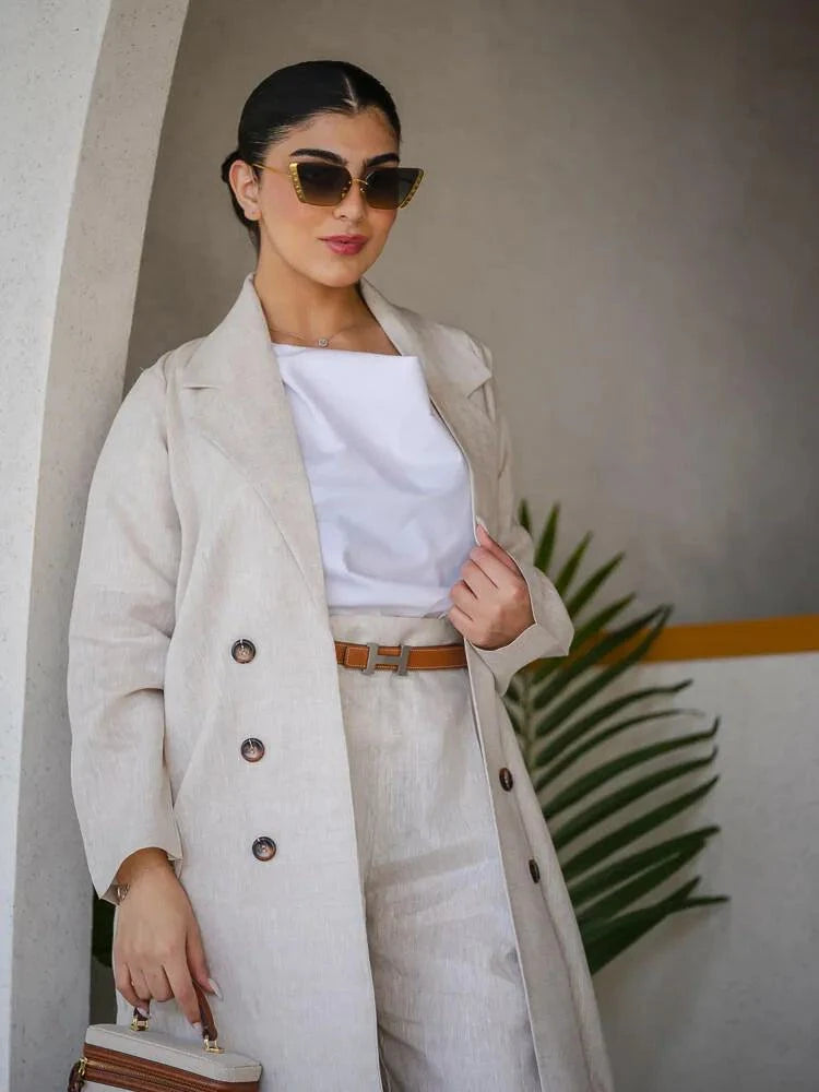 Woman wearing Whisper Beige linen abaya with pleated chiffon panels and double-breasted buttons, styled with a white top, beige skirt, and heels.