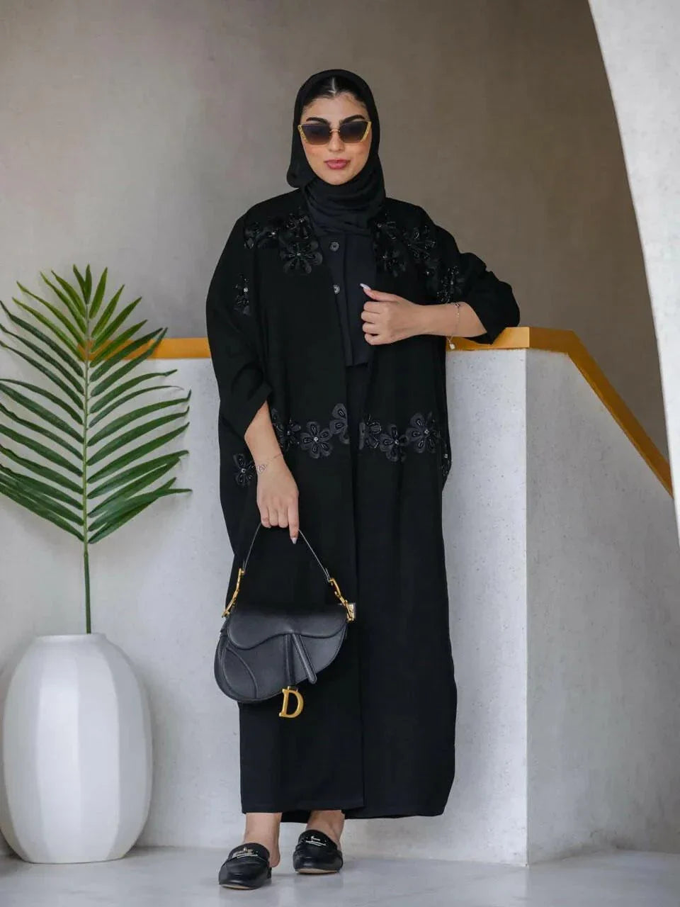 Woman wearing a black Elegant Abaya with delicate embroidery and lace, accessorized with sunglasses and a black handbag, standing indoors near a white planter with a green plant.
