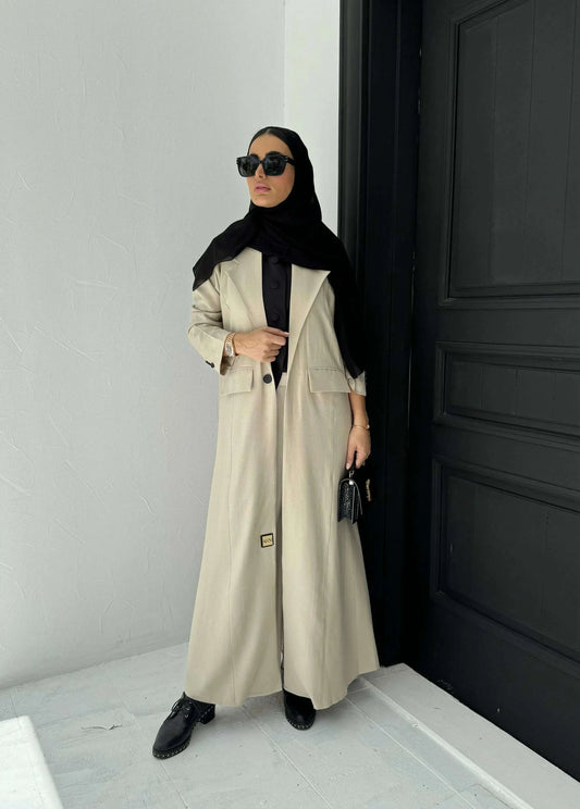 Woman wearing beige linen blazer-style abaya with lapel collar, single-button closure, front flap pockets, and turned-up cuffs.