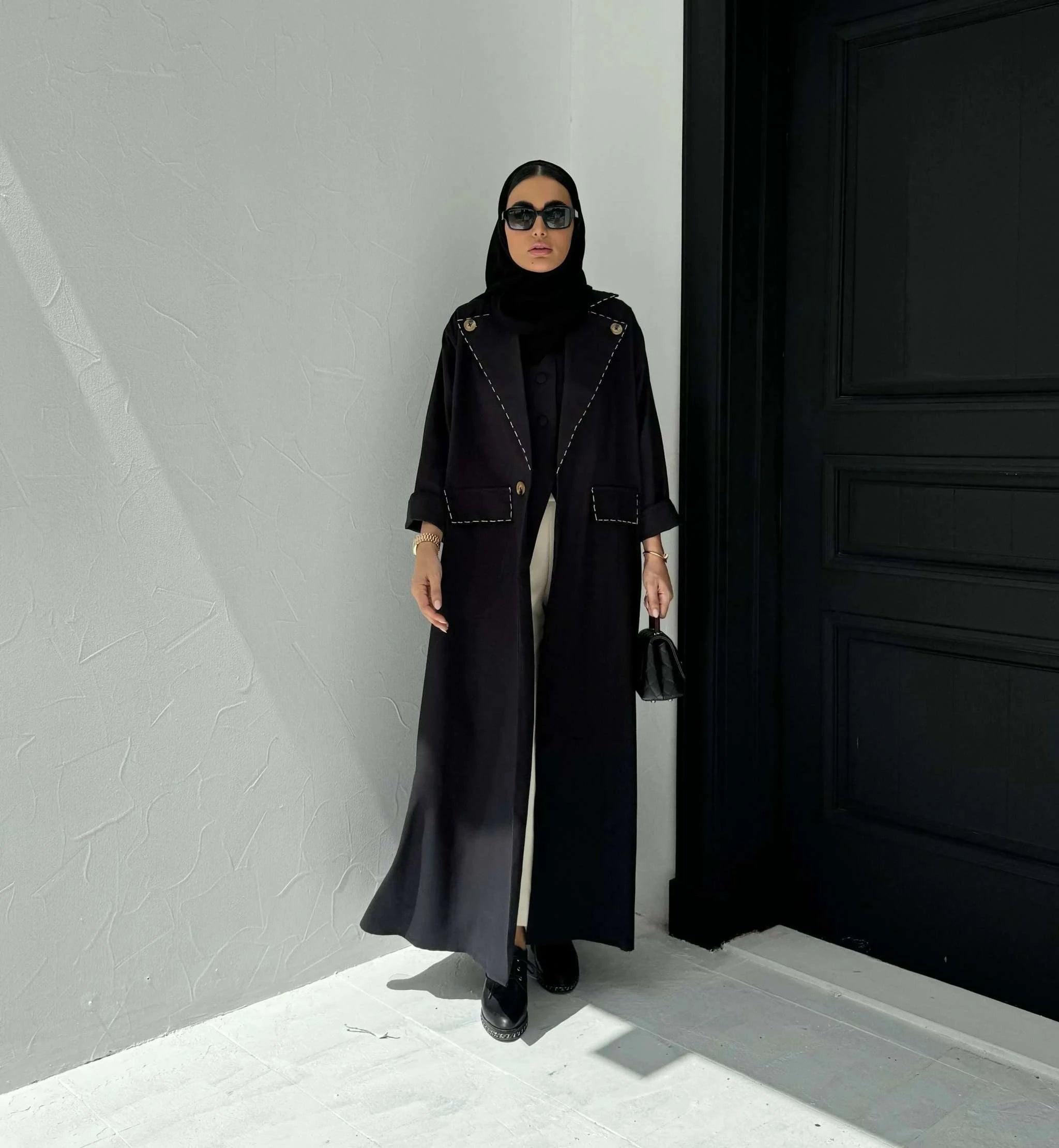 Woman wearing a long black blazer abaya with gold buttons, sharp lapels, rolled cuffs, and false flap pockets.