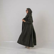 Woman wearing an olive green lightweight abaya made of cobra silk with soft cloche cut and back pleats, accessorized with sunglasses and a handbag.