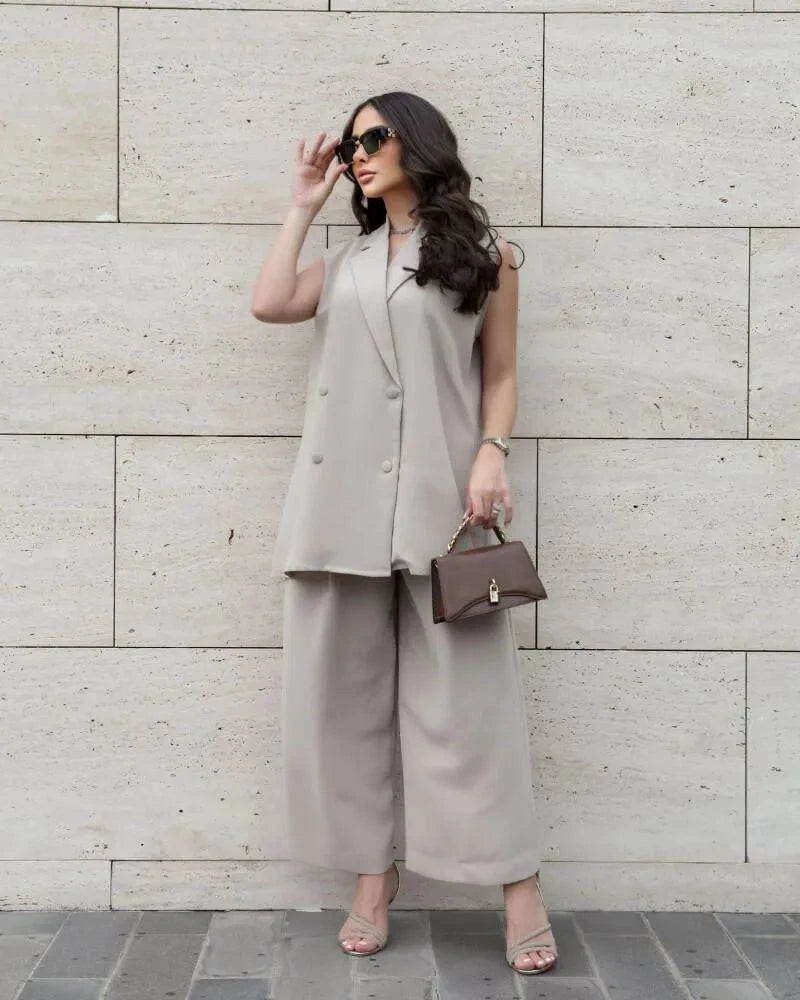 Woman wearing a stylish light beige sleeveless double-breasted vest and wide-leg pants inner set with sunglasses and handbag against a stone wall background