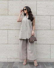 Woman wearing a stylish light beige sleeveless double-breasted vest and wide-leg pants inner set with sunglasses and handbag against a stone wall background