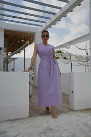Woman wearing a soft purple orchid linen dress with a relaxed silhouette and tied waist, standing outdoors under a pergola.