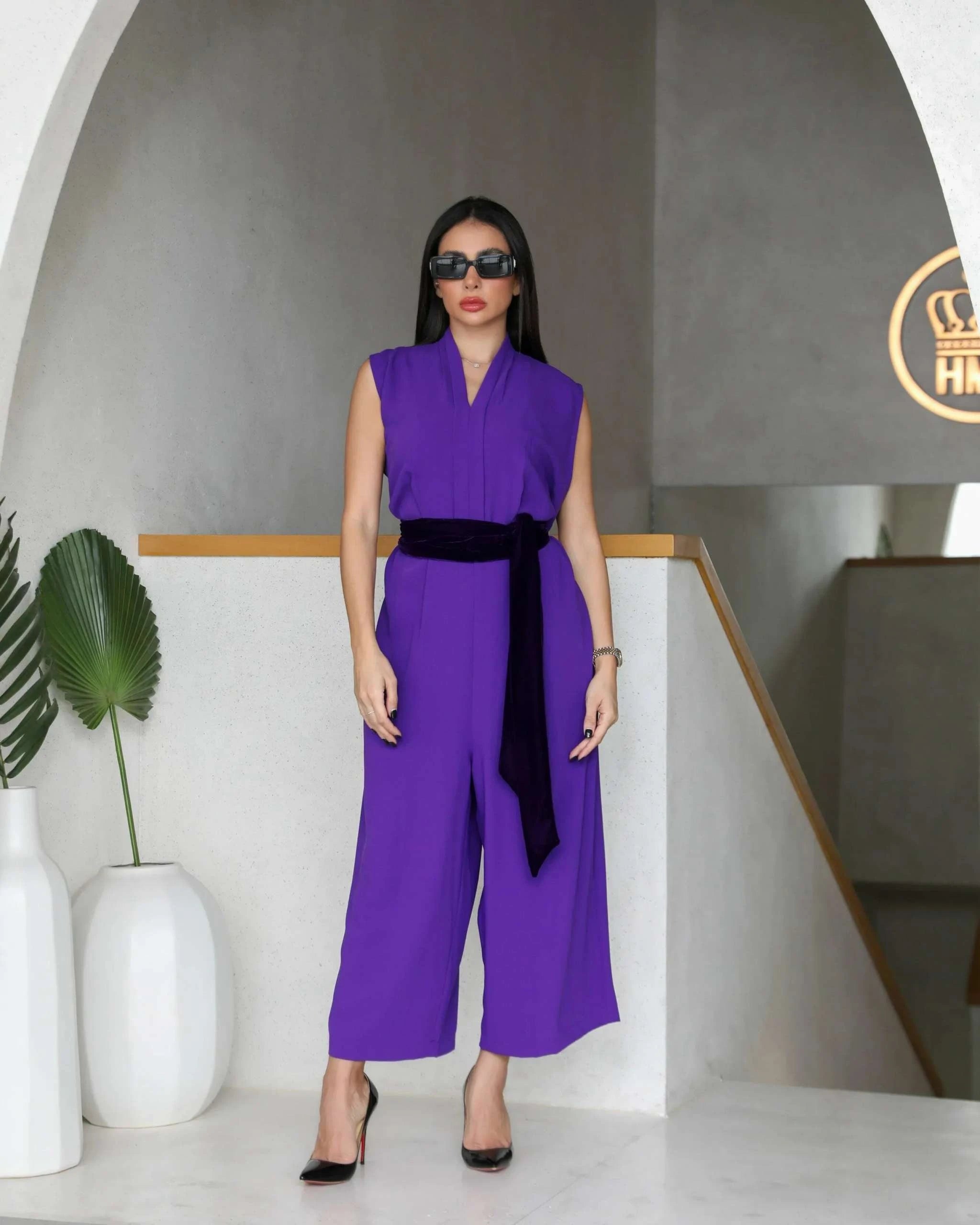 Woman wearing a dark purple crepe sleeveless jumpsuit with a black belt and black heels in a modern indoor setting