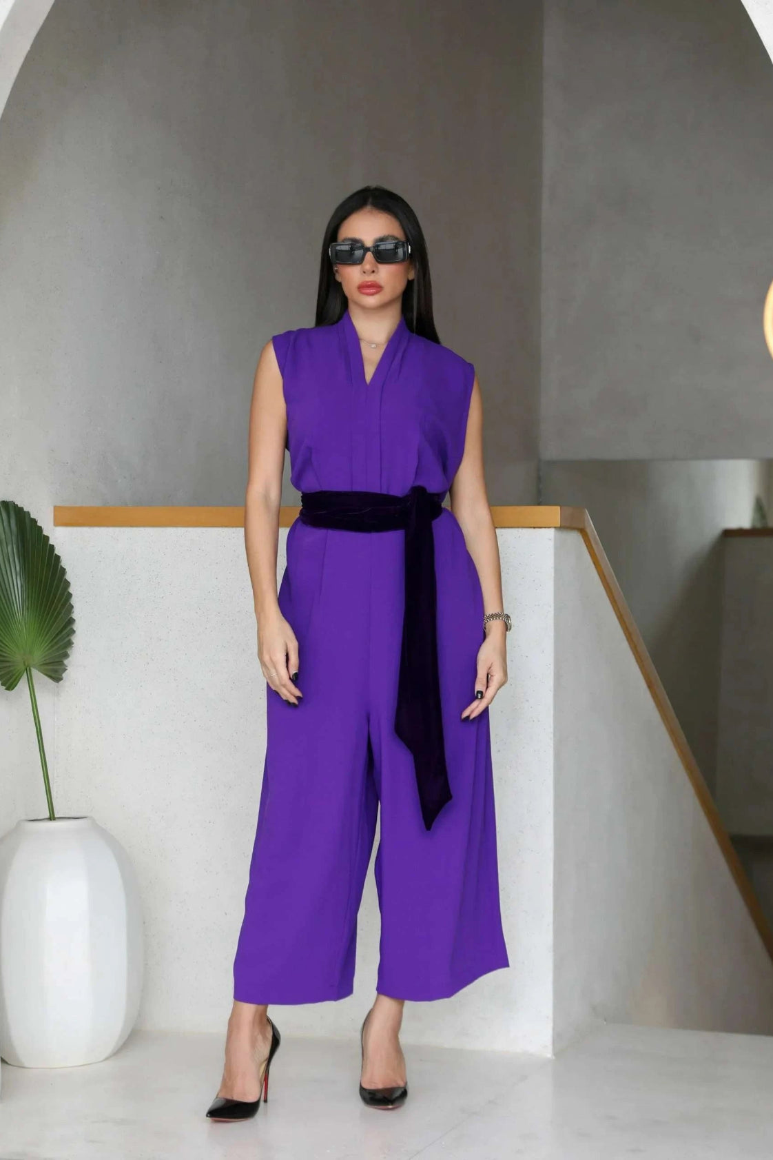 Woman wearing a dark purple crepe sleeveless jumpsuit with a black belt and black heels in a modern indoor setting