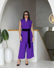 Woman wearing a dark purple crepe sleeveless jumpsuit with a black belt and black heels in a modern indoor setting