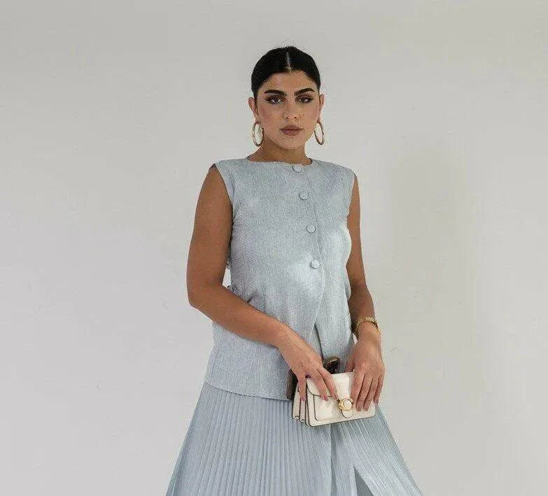 Woman wearing sleeveless sky blue linen vest with buttons, holding a small beige purse against a plain background