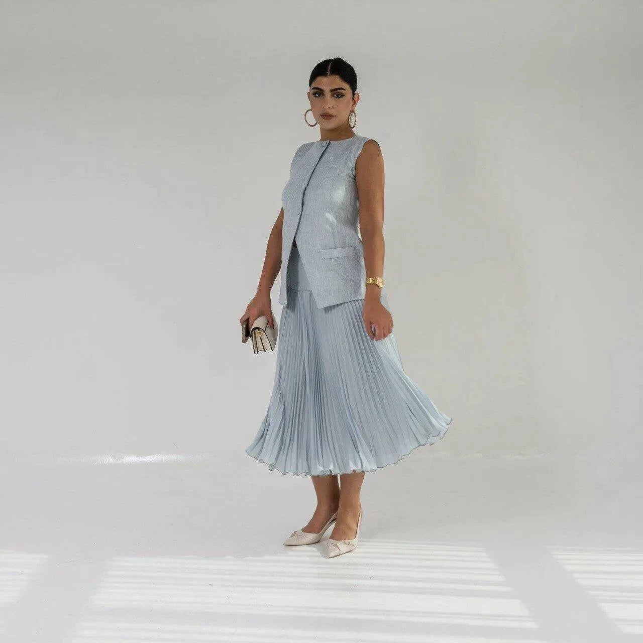 Woman wearing sleeveless sky blue linen vest with buttons, holding a small beige purse against a plain background