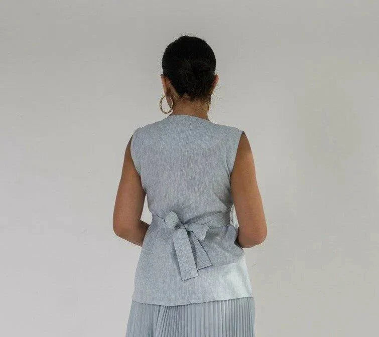 Woman wearing sleeveless sky blue linen vest with buttons, holding a small beige purse against a plain background