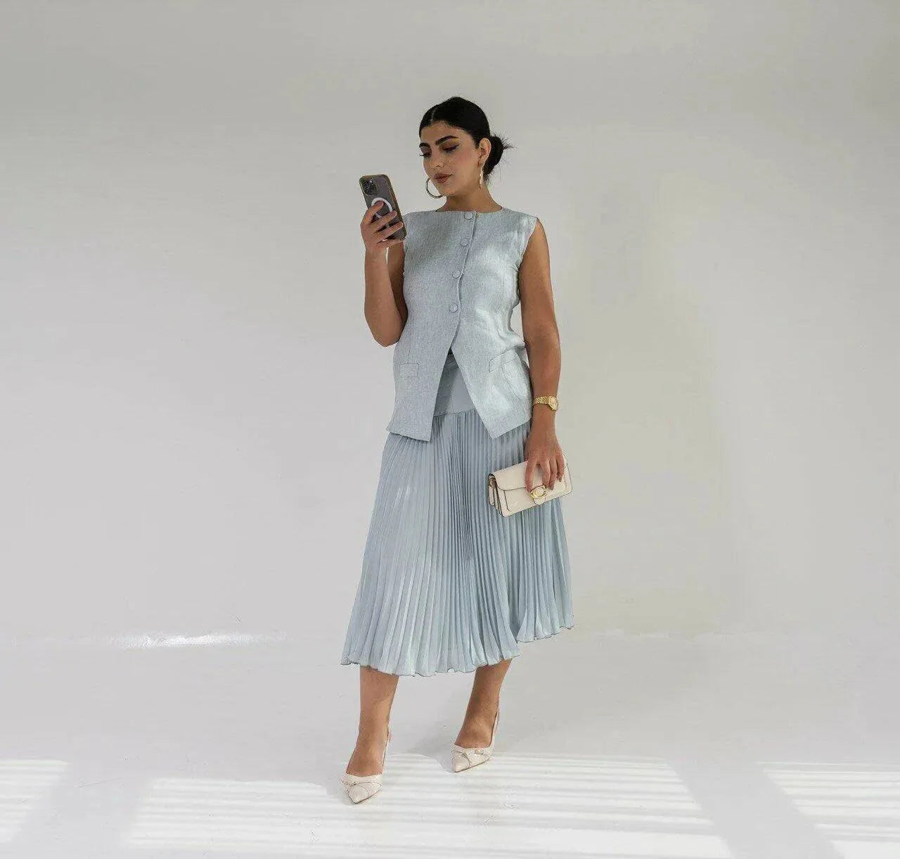 Woman wearing a flowy sky blue chiffon skirt with a sleeveless gray top and beige heels holding a phone and clutch purse
