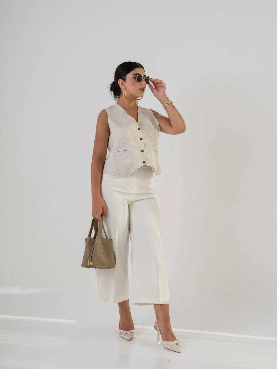 Woman wearing tailored white crepe pants with sleeveless beige vest and holding a taupe handbag
