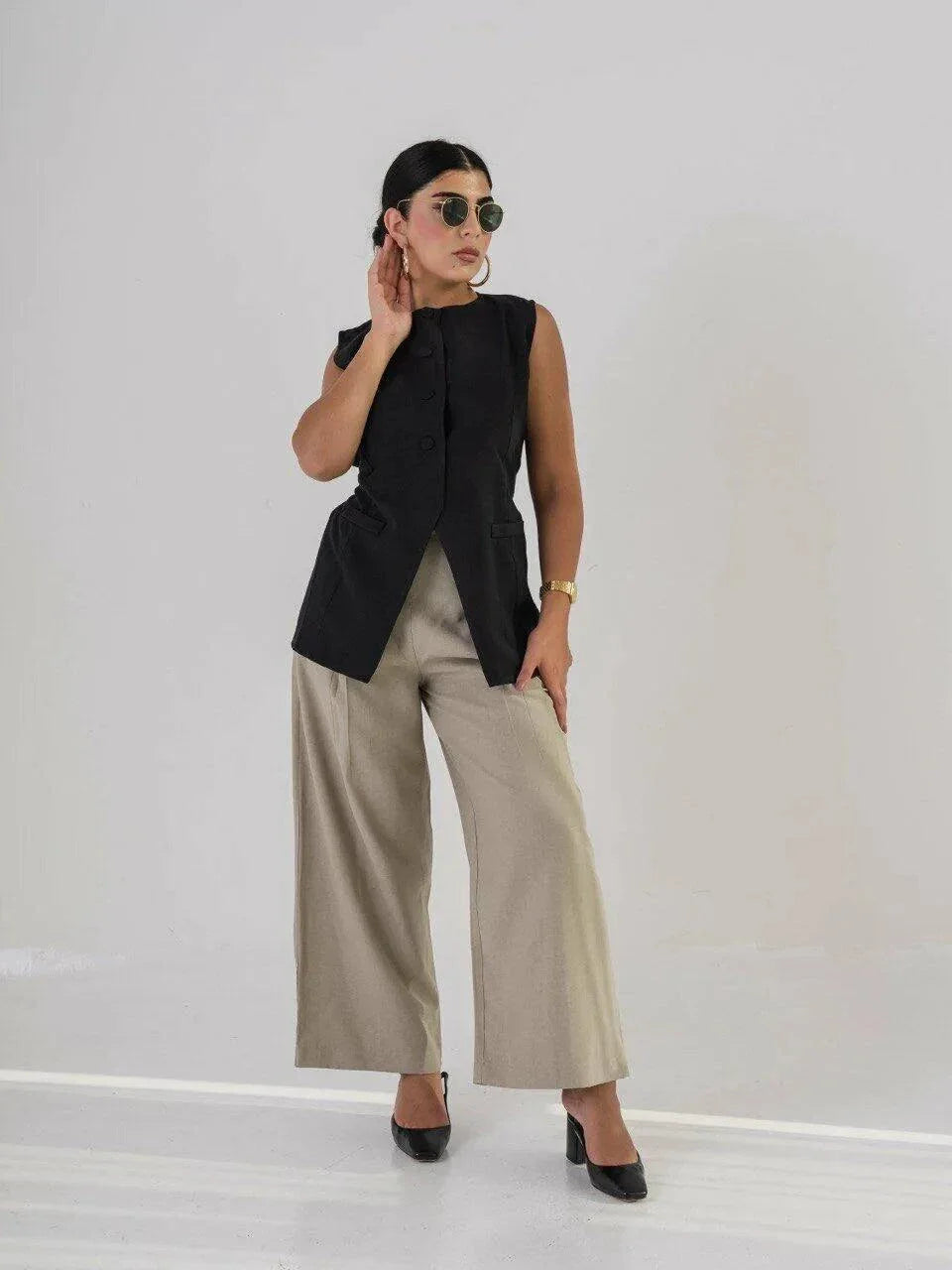 Woman wearing long black crepe vest with beige wide-leg pants and sunglasses on white background