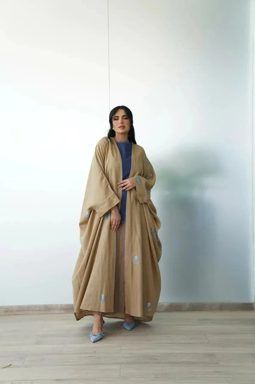 Woman wearing camel-toned linen bisht with blue geometric embroidery over blue dress indoors