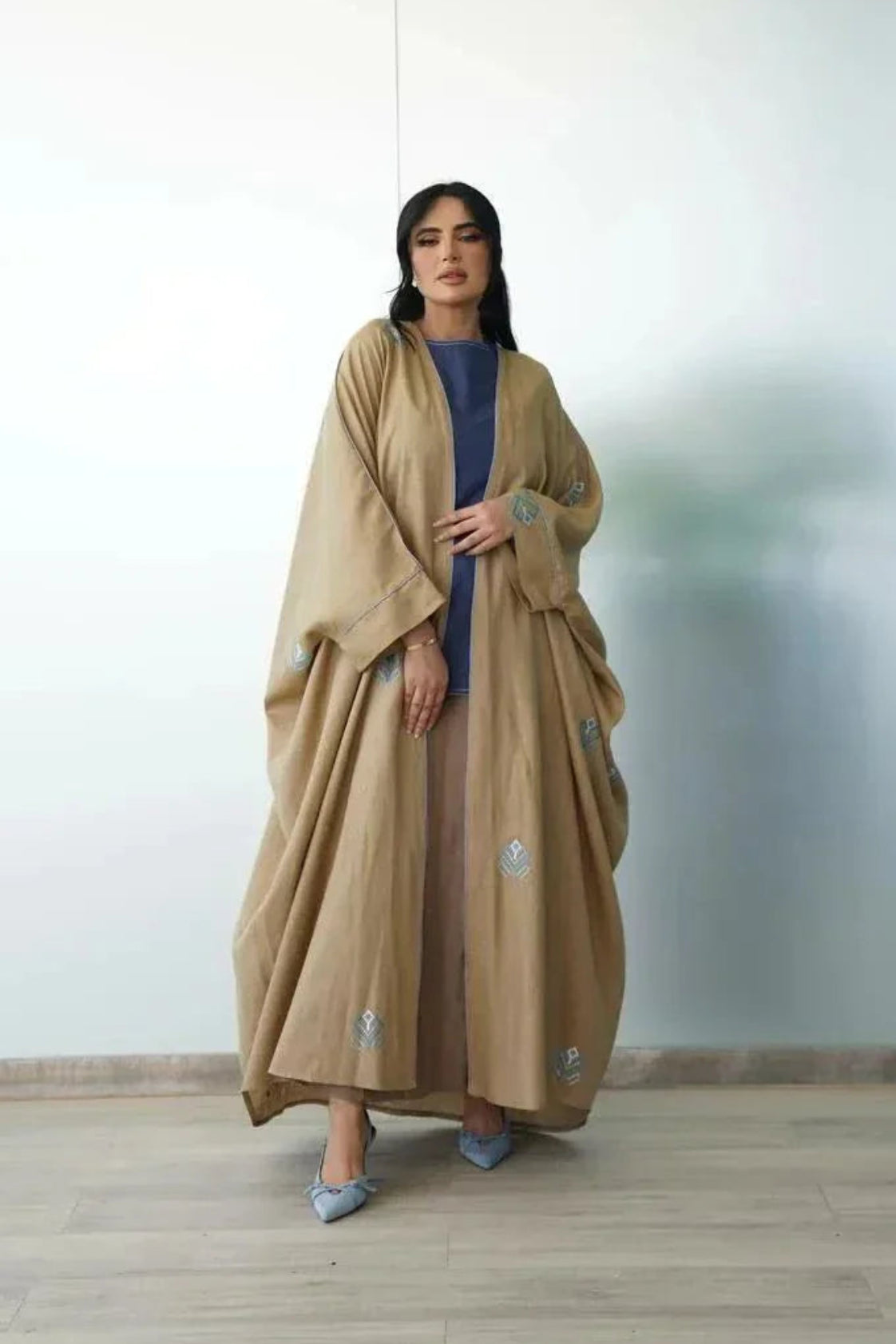 Woman wearing camel-toned linen bisht with blue geometric embroidery over blue dress indoors