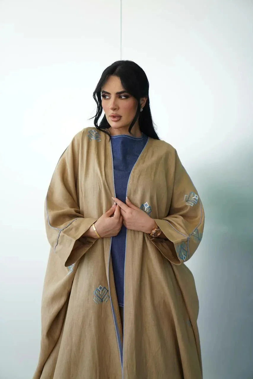 Woman wearing a camel-toned linen bisht with soft blue geometric embroidery and wide sleeves.