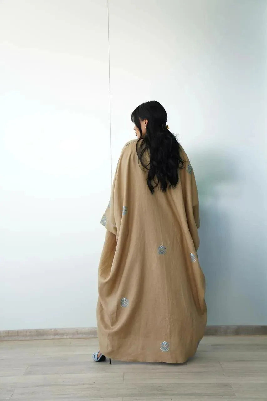 Woman wearing a camel-toned linen bisht with soft blue geometric embroidery and wide sleeves.