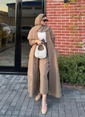 Woman wearing a beige abaya with wide flowy design, paired with matching pants, hijab, sunglasses, and carrying a white handbag outdoors.